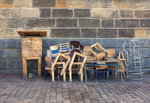 The image displays a collection of discarded wooden chairs stacked and leaning against a dark brick wall on a cobblestone pavement. The chairs vary in design and finish, with some showing natural wood tones, while others are painted or have a painted finish. To the left, there is a small wooden storage unit with a lid, and on the right side, a metallic stepladder is positioned beside the chairs. The scene appears to be outdoors, possibly on a pavement in Peckham, with the background consisting of dark, weathered brickwork, suggesting a waste or recycling area managed by Commercial Waste Peckham as part of their rubbish removal services. The lighting indicates daylight conditions, and the overall appearance is of unwanted furniture items awaiting collection or disposal.