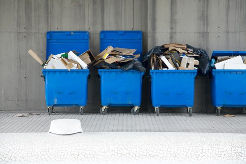 The image shows a row of four large, bright blue wheelie bins placed on a paved sidewalk against a grey concrete wall. The bins are filled with assorted waste materials, including flattened cardboard boxes, paper, and miscellaneous packaging, with some bits spilling over the edges. The second and third bins from the left appear particularly full, with cardboard and paper protruding from the open top. The first bin on the left has its lid partially open, revealing similar contents, while the fourth bin to the far right is also filled with waste, some of which is covered with a black plastic bag. In the foreground, there is a white plastic container lying on the ground, slightly away from the bins. The setting appears to be an outdoor space, possibly part of a commercial or industrial area, suitable for rubbish collection services in Peckham, serving local businesses requiring regular waste removal handled by companies such as Commercial Waste Peckham.