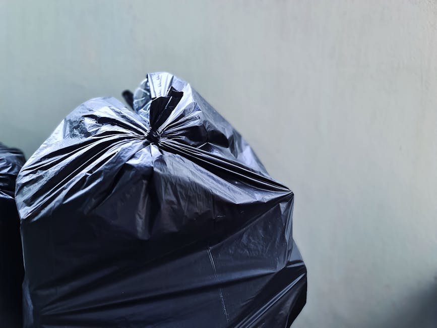 A worker wearing a blue uniform and a high-visibility orange vest is positioned on the right side of the image, emptying a blue wheelie bin into the rear compartment of a white garbage collection truc